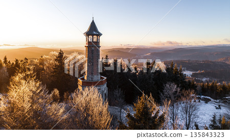 The Stepanka lookout tower stands tall in the Jizera Mountains as the sun sets. The landscape features trees and light snow, creating a clear view of the surrounding hills and valleys. The Stepanka lookout tower stands tall in the Jizera Mountains as the sun sets. The landscape features trees and light snow, creating a clear view of the surrounding hills and valleys. 135843323