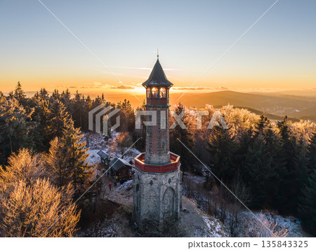 Stepanka lookout tower stands tall in the Jizera Mountains during winter. Visitors enjoy a sunset view over the landscape and surrounding trees in this serene mountain setting. 135843325