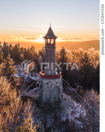 Visitors gather at Stepanka lookout tower in the Jizera Mountains during sunset. The scene features frost-covered trees and a beautiful sky, creating a striking winter atmosphere. Visitors gather at Stepanka lookout tower in the Jizera Mountains during sunset. The scene features frost-covered trees and a beautiful sky, creating a striking winter atmosphere. 135843326
