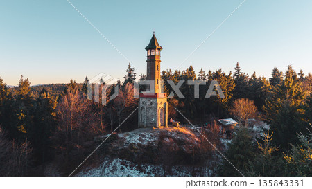 Stepanka lookout tower stands tall in the Jizera Mountains surrounded by snow-covered trees. The sunset casts warm light on the tower creating a beautiful scene. Stepanka lookout tower stands tall in the Jizera Mountains surrounded by snow-covered trees. The sunset casts warm light on the tower creating a beautiful scene. 135843331