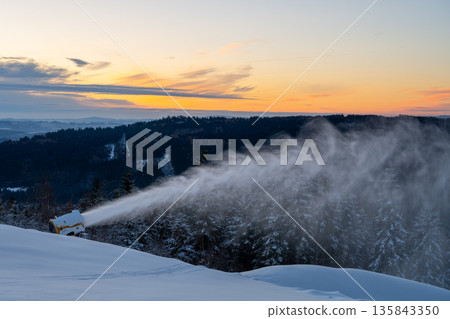 Snow cannon operates on a mountain slope, creating artificial snow for skiing. The sun sets in the background, adding color to the winter landscape. Trees are covered in snow. Snow cannon operates on a mountain slope, creating artificial snow for skiing. The sun sets in the background, adding color to the winter landscape. Trees are covered in snow. 135843350