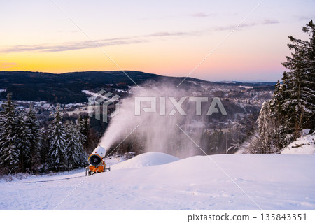 A snow cannon produces snow on a ski slope at sunset. Snow covers the ground, and mountains stretch in the background. Skiers will enjoy this area for winter sports soon. A snow cannon produces snow on a ski slope at sunset. Snow covers the ground, and mountains stretch in the background. Skiers will enjoy this area for winter sports soon. 135843351