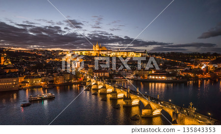 The scene captures Prague Castle and Charles Bridge illuminated at night. The river Vltava reflects lights from the city. It shows a vibrant skyline and evening activity. 135843353
