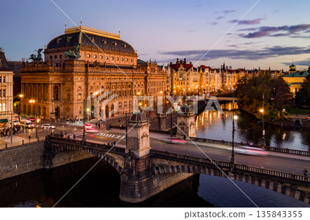 Aerial view of the National Theatre in Prague at evening. The theatre is illuminated against a darkening sky, with the Vltava River and embankment visible. Traffic moves across the bridge. 135843355
