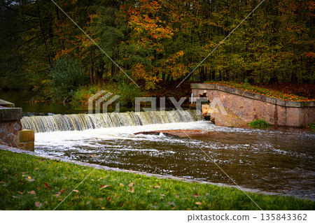 A beautiful waterfall flows gently through Granny Walley in Ratiborice. The area is surrounded by colorful autumn trees, creating a peaceful rural scene. Visitors enjoy the serene environment. 135843362