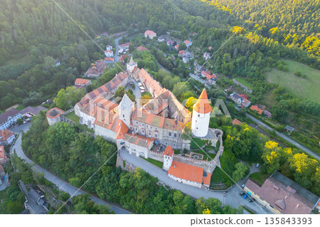 Krivoklat Castle stands majestically in central Bohemia, surrounded by lush greenery. The warm colors of sunset illuminate the medieval architecture, creating a picturesque scene. Krivoklat Castle stands majestically in central Bohemia, surrounded by lush greenery. The warm colors of sunset illuminate the medieval architecture, creating a picturesque scene. 135843393