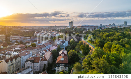 Morning sunlight bathes the Vysehrad Basilica of St. Peter and St. Paul, highlighting the stunning architecture against the backdrop of Prague's skyline and lush greenery. 135843398