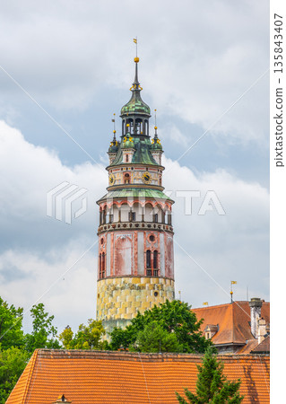 The castle tower stands tall among the roofs of Cesky Krumlov. This notable landmark attracts visitors with its history and architecture. Green trees surround the base. 135843407