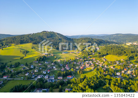 Tanvaldsky Spicak stands majestically in the Jizera Mountains, showcasing lush green hills and charming villages below. Warm sunlight accentuates the natural landscape, inviting exploration. Tanvaldsky Spicak stands majestically in the Jizera Mountains, showcasing lush green hills and charming villages below. Warm sunlight accentuates the natural landscape, inviting exploration. 135843422