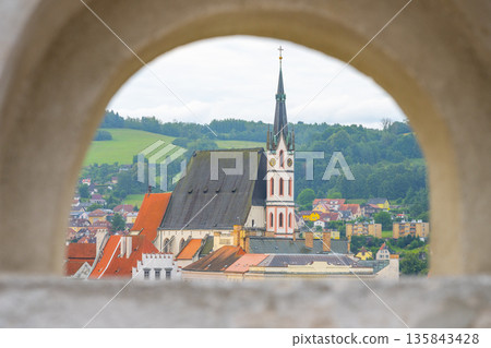 Saint Vitus Church stands in Cesky Krumlov under cloudy skies. The view captures the church's tower and surrounding colorful buildings in the UNESCO-listed town, showcasing summer life. 135843428