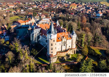 Zleby Castle is shown from the air, surrounded by trees and a small village. The castle has distinctive towers and a red roof. It is located in the countryside. Zleby Castle is shown from the air, surrounded by trees and a small village. The castle has distinctive towers and a red roof. It is located in the countryside. 135843626