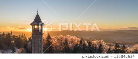 Stepanka lookout tower stands tall in the Jizera Mountains during winter. The sunset casts a warm glow over the snowy landscape, showcasing nature's beauty and peaceful surroundings. 135843634