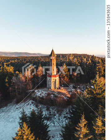 Stepanka lookout tower stands tall in the Jizera Mountains during winter. Sunlight shines on the tower surrounded by snow-covered trees and mountains in the background. Stepanka lookout tower stands tall in the Jizera Mountains during winter. Sunlight shines on the tower surrounded by snow-covered trees and mountains in the background. 135843635