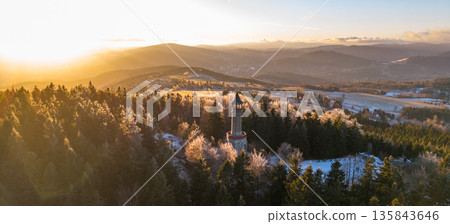 Stepanka lookout tower stands tall in the Jizera Mountains as the sun sets, casting golden light over snowy trees and the landscape below. Visitors enjoy the winter scenery. Stepanka lookout tower stands tall in the Jizera Mountains as the sun sets, casting golden light over snowy trees and the landscape below. Visitors enjoy the winter scenery. 135843646