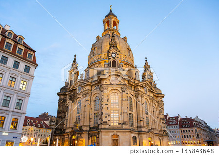 Dresden Frauenkirche features a large dome and ornate architecture. The church is surrounded by buildings and a clear sky during the evening. People visit the site during Christmas time. 135843648