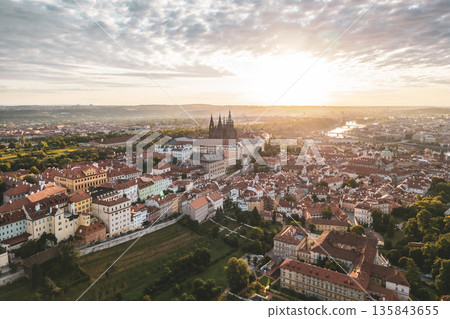 Glowing sunlight casts a warm hue over Prague Castle as it rises above the historic city. The vibrant rooftops and lush greenery create a picturesque view at dawn, awakening the landscape. 135843655