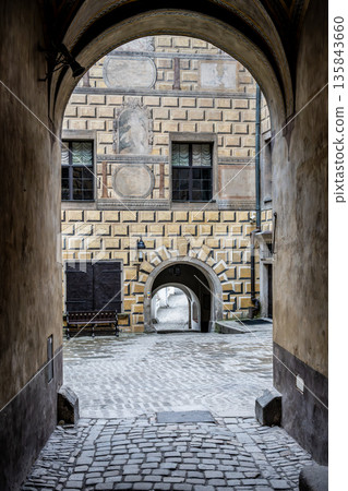 A view from a stone archway showing the cobblestone street and surrounding buildings in Cesky Krumlov, Czechia. The summer day adds life to this historic location. A view from a stone archway showing the cobblestone street and surrounding buildings in Cesky Krumlov, Czechia. The summer day adds life to this historic location. 135843660