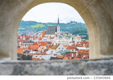 Visitors can see the rooftops of Cesky Krumlov through a stone arch. The town includes buildings with red roofs and a tall church steeple in the background. It is a summer day. 135843668