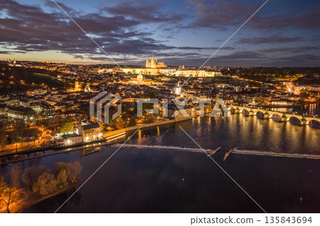 An aerial view of Prague at night. The Vltava River flows beneath the Charles Bridge while the castle is illuminated in the distance. Buildings and streets shine with lights. An aerial view of Prague at night. The Vltava River flows beneath the Charles Bridge while the castle is illuminated in the distance. Buildings and streets shine with lights. 135843694