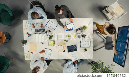 Top down view of business people consulting on finance and budgeting during a corporate meeting. Staff analyze documents, diagrams and reports to align goals and planning strategies. Top down view of business people consulting on finance and budgeting during a corporate meeting. Staff analyze documents, diagrams and reports to align goals and planning strategies. 135844382
