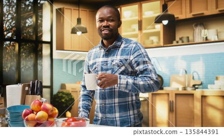 Portrait of cheerful young guy holding a cup of fresh coffee at counter, delicious pastry on a plate. Scent of toast and coffee fills the air during morning routine with homemade meal. Portrait of cheerful young guy holding a cup of fresh coffee at counter, delicious pastry on a plate. Scent of toast and coffee fills the air during morning routine with homemade meal. 135844391