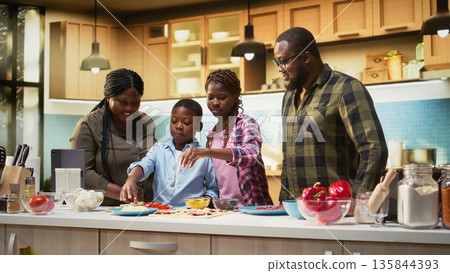 Smiling children help parents prepare pizza by sprinkling grated cheese in the kitchen, family cooking activity during the weekend. Cozy moment of teamwork and meal preparation. Camera A. 135844393