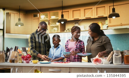 Parents and children having fun in the kitchen cooking homemade pizza, layering ingredients like pepperoni and sauce while enjoying laughter and teamwork. Cozy weekend activity. Camera A. 135844395