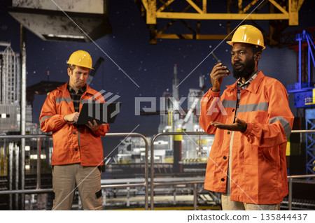 Black drilling rig employee speaking through walkie talkie with colleagues, monitoring oil extraction systems for efficiency. Trying to minimize carbon footprint levels on platform. Black drilling rig employee speaking through walkie talkie with colleagues, monitoring oil extraction systems for efficiency. Trying to minimize carbon footprint levels on platform. 135844437