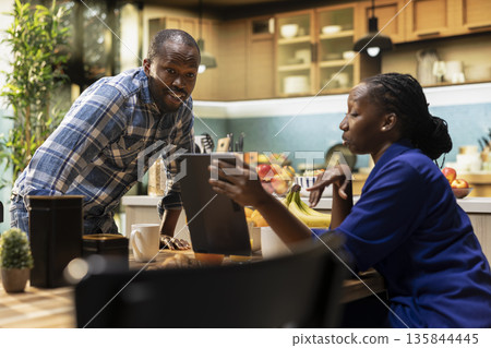 Black cute lovers laughing at videos on tablet enjoying breakfast in the kitchen. Couple checking social media and sipping coffee, both relaxed and smiling during morning routine together. Black cute lovers laughing at videos on tablet enjoying breakfast in the kitchen. Couple checking social media and sipping coffee, both relaxed and smiling during morning routine together. 135844445