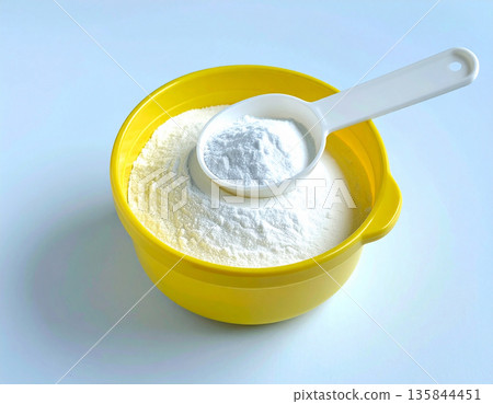 Powdered agent for washing. Pile of powdered detergent in yellow plastic container with a measuring spoon on a white background. Home washing and cleaning. 135844451
