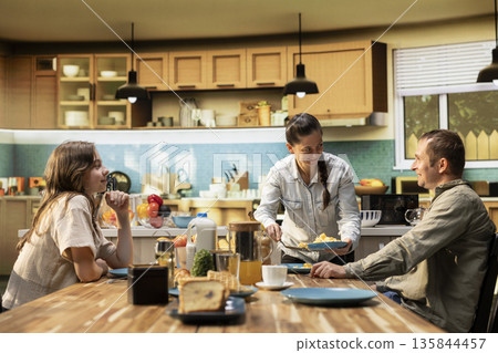 Family sharing scrambled eggs and pastry for breakfast at home, parents serving omelettes and toast with little boy and pre-teen girl. Warmth, relaxation and joyful togetherness. Family sharing scrambled eggs and pastry for breakfast at home, parents serving omelettes and toast with little boy and pre-teen girl. Warmth, relaxation and joyful togetherness. 135844457