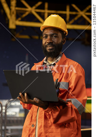 African american engineer in hard hat performing analysis on laptop from offshore platform, drilling operations align with efficient oil and fuel extraction. Marine habitat contamination. 135844467