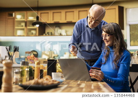 Old retired people shopping online for food with their credit card, reviewing grocery shopping items and healthy eating produce in app cart on laptop. Convenient home delivery options. 135844468