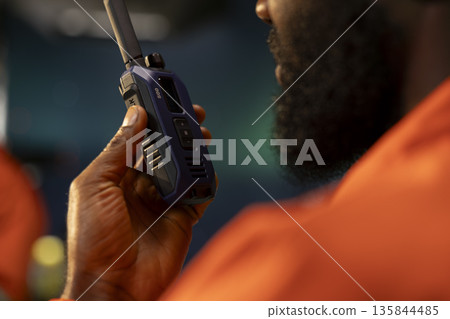 Close up of black engineer speaking via walkie talkie with colleagues from drilling rig, monitoring oil extraction activity for efficiency. Trying to minimize carbon footprint levels on platform. 135844485