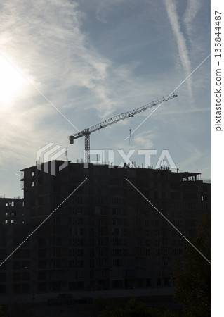Tower crane lifting heavy materials for construction of high rising building during city expansion. Heavy machinery used for apartment complex construction project over blue sky Tower crane lifting heavy materials for construction of high rising building during city expansion. Heavy machinery used for apartment complex construction project over blue sky 135844487