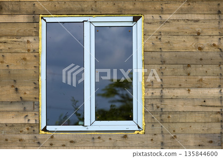 Close-up detail of new narrow plastic vinyl window installed in house wall of natural wooden planks. 135844600