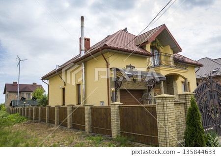 New yellow two-story residential cottage with shingle roof, front porch, plastic windows, high chimneys, balconies on blue sky background. Real estate property, architecture and prosperity concept. 135844633
