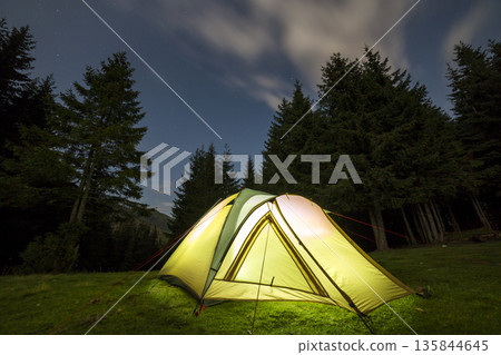 Summer camping at night. Illuminated tourist tent on green clearing on distant mountain background. 135844645