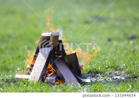 Glowing bonfire on nature. Burning wooden planks outside on summer day. Bright orange flames, light smoke and dark ashes on green grass on blurred green background. Tourism and camping concept. 135844654