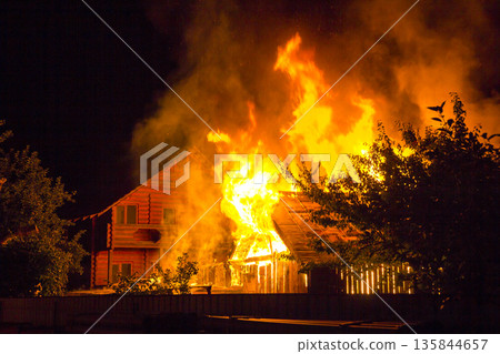 Burning wooden house at night. Bright orange flames and dense smoke from under the tiled roof on dark sky, trees silhouettes and residential neighbor cottage background. Disaster and danger concept. 135844657