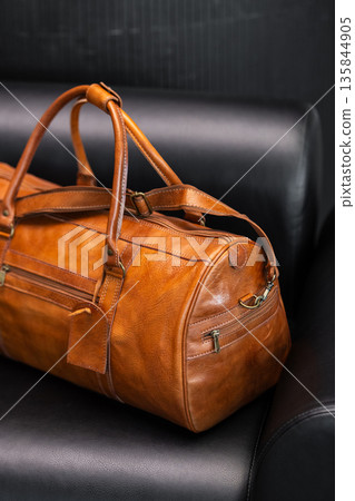 Premium tan leather duffel bag on a black leather sofa in a modern minimalist interior Premium tan leather duffel bag on a black leather sofa in a modern minimalist interior 135844905