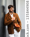 Close up of a stylish man in a brown suede jacket holding a tan leather wristlet clutch bag 135844908