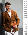 Close up of a stylish man in a brown suede jacket holding a tan leather wristlet clutch bag 135844909