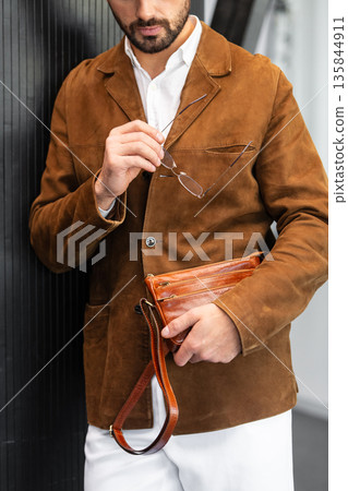 Close up of a stylish man in a brown suede jacket holding a tan leather wristlet clutch bag Close up of a stylish man in a brown suede jacket holding a tan leather wristlet clutch bag 135844911