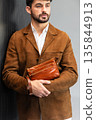 Close up of a stylish man in a brown suede jacket holding a tan leather wristlet clutch bag 135844913
