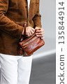 Close up of a stylish man in a brown suede jacket holding a tan leather wristlet clutch bag 135844914