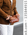 Close up of a stylish man in a brown suede jacket holding a tan leather wristlet clutch bag 135844915