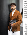 Stylish man in a brown suede blazer and white trousers leaning against a wall holding a small dark leather crossbody bag 135844918