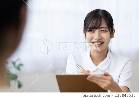 A smiling young woman in a white coat talking to a patient while filling out a medical questionnaire A smiling young woman in a white coat talking to a patient while filling out a medical questionnaire 135845533