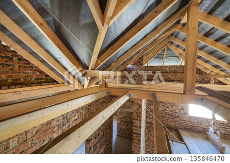 Attic of a building under construction with wooden beams of a roof structure and brick walls. 135846470